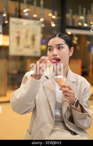 A cheerful young Southeast Asian Thai woman from the Isan region sitting inside a modern shopping mall while putting a small red spoon to her mouth to Stock Photo