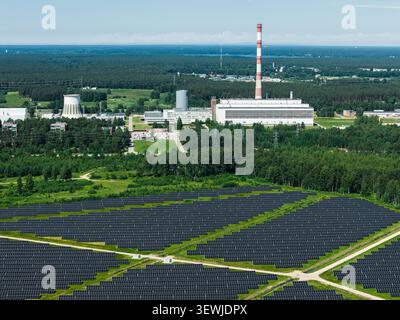 Aerial view of a large solar farm adjacent to a power plant with a tall chimney, set amidst expansive green forests in Riga, Latvia. Stock Photo