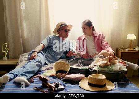 Young adult couple preparing for vacation, packing suitcase on bed and discussing travel plans, representing trip planning and partnership. Wide shot, ideal for tourism ads and booking campaigns Stock Photo