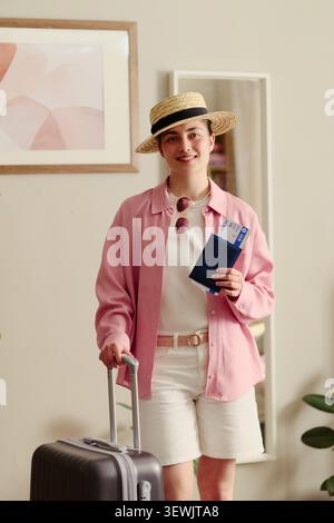 Portrait of young adult Caucasian woman holding passport and boarding pass, pulling suitcase and preparing for vacation travel. Ideal for tourism marketing, trip planning, vertical Stock Photo