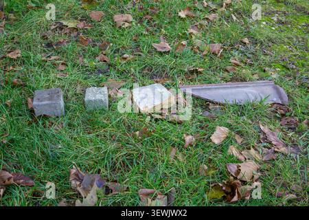Two different blocks or cobblestones next to a piece of wall and a broken piece of grey clay on green grass with brown leaves, group of stones in back Stock Photo