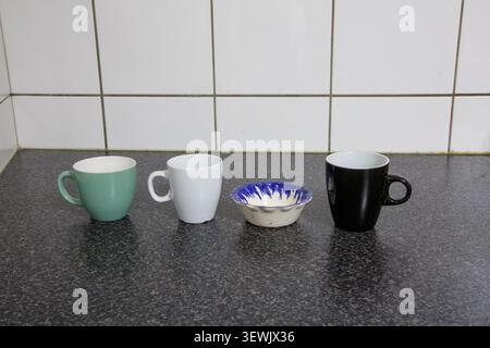 Saucer among three wide-mouthed ceramic cups of different shapes and sizes on black kitchen counter, white tiled wall, household utensils for preparin Stock Photo