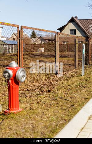 A bright red vintage fire hydrant stands on a grassy roadside next to a wire fence in a quiet residential neighborhood. Stock Photo
