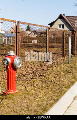 A bright red vintage fire hydrant stands on a grassy roadside next to a wire fence in a quiet residential neighborhood. Stock Photo