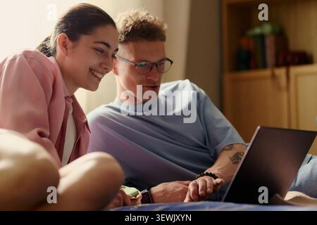 Young adult couple using laptop together at home, browsing online content or booking vacation. Ideal for remote lifestyle, digital services, e learning, medium shot Stock Photo