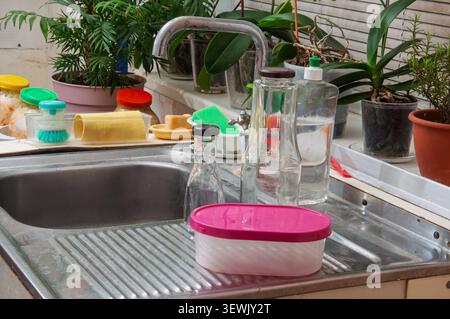 A cluttered kitchen sink area features various plants, bottles, sponges, and food storage containers on the countertop Stock Photo