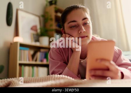 Young adult Caucasian woman lying on bed and using smartphone at home, browsing social media or messages. Suitable for digital lifestyle, mobile app, communication marketing Stock Photo