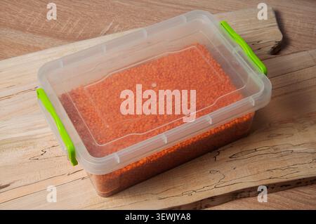 Red lentils stored in a clear plastic container with green clasps on a wooden surface Stock Photo