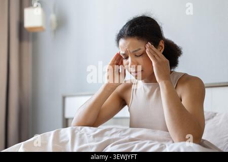 Black woman sitting in bed and touching her temples with closed eyes Stock Photo