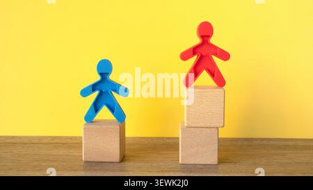 A blue plastic figure standing on a low wooden block beside a red plastic figure standing on a higher stack of blocks on a yellow background, demonstr Stock Photo