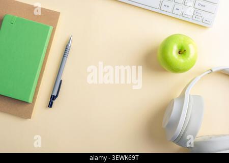 Minimalist workspace with notebook, pen, keyboard, headphones and green apple on a beige desk. Top view, flat lay, copy space. Stock Photo