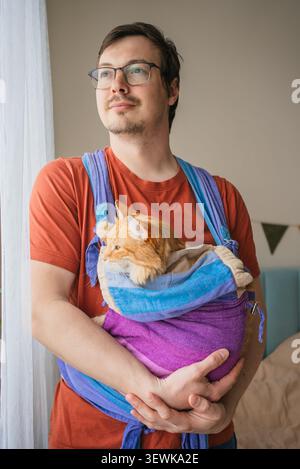 Man holding a fluffy ginger cat in a vivid purple and blue sling carrier, standing indoors near a window with soft light, embracing comfort and companionship Stock Photo