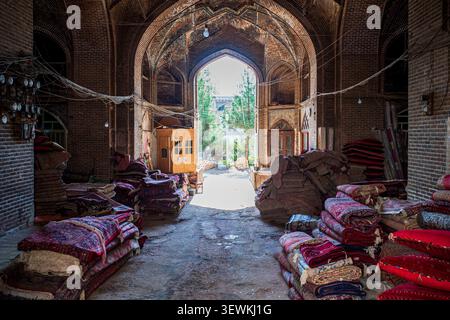 Interior of an ancient brick covered market. Large stacks of traditional Persian carpets with red and blue geometric patterns lie on the floor, while Stock Photo