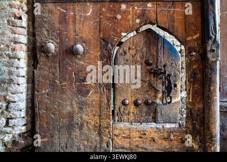 Close-up shot of an old, time-worn solid wooden door. The center features a smaller arched wicket door secured with a heavy iron chain and decorated w Stock Photo
