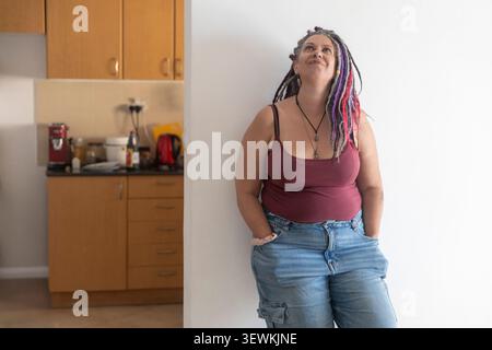 A middle-aged woman with colorful dreadlocks stands leaning against a white wall in her kitchen, looking up thoughtfully with a smile. Stock Photo