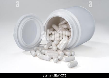White plastic pill bottle tipped over on its side with numerous clear medicine capsules filled with white powder spilling out onto a clean white backg Stock Photo