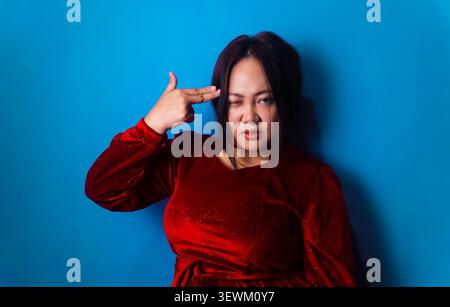Young Asian woman wearing a red velvet dress makes a finger gun gesture against her temple while winking at the camera against a blue background Stock Photo