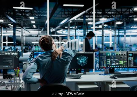 Software developer stretches at a workstation in a server room to relieve back fatigue after long technical work. Stock Photo