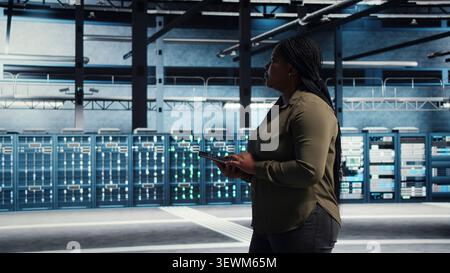 Data center technician monitors operational metrics on tablet inside a server room to improve infrastructure performance. Stock Photo