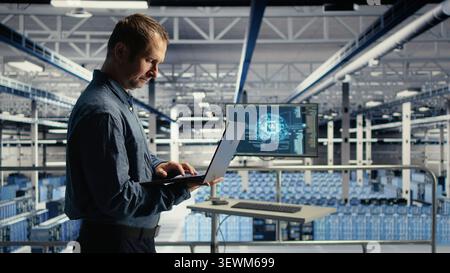Data center technician monitors operational metrics on laptop inside a server room to improve infrastructure performance. Stock Photo