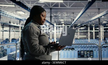 African american IT professional uses a laptop in a server room to review test results and confirm reliable system operation. Stock Photo