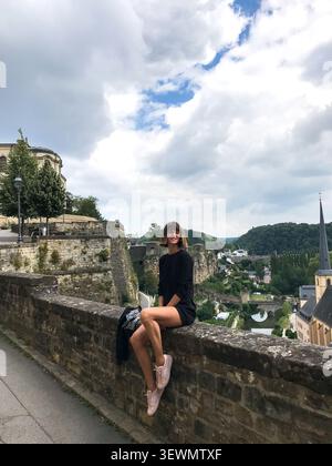 A beautiful young woman sits on the edge of a viewpoint overlooking the historic city of Luxembourg. The female model enjoys the panoramic cityscape. Stock Photo