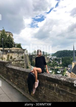 A beautiful young woman sits on the edge of a viewpoint overlooking the historic city of Luxembourg. The female model enjoys the panoramic cityscape. Stock Photo
