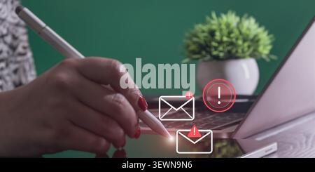 Close-up of hand with digital pen touching a screen with floating urgent email notification graphics, laptop in background. Stock Photo