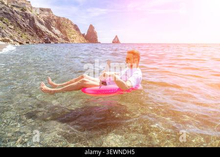 Freelancer remote vacation man works on a tablet while floating in a pink inflatable ring on clear sea water under bright sunshine Stock Photo