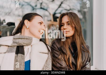 Friends talking outdoors two young women in autumn coats sharing a conversation by a building on a sunny day Stock Photo