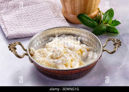 Soft white yogurt in hammered copper bowl with vintage handles. Fresh mint leaves wooden mortar in background. Pastel cloth adds gentle texture Stock Photo
