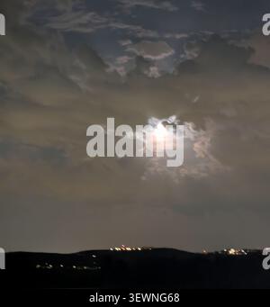 Full moon shining brightly through dark, dramatic storm clouds over a distant city skyline at night. Stock Photo