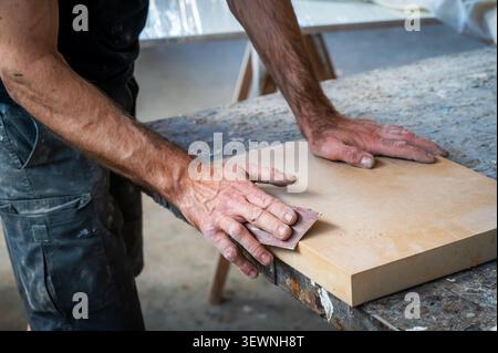 A carpenter manually sands the edge of a wood panel using sandpaper in a woodworking workshop, showcasing traditional craftsmanship and attention to d Stock Photo