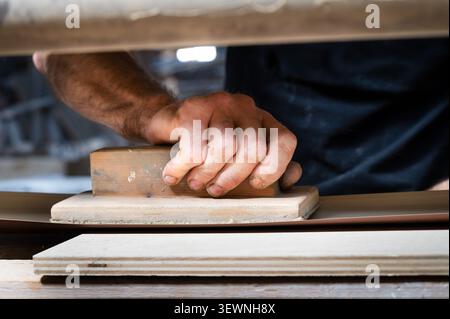 Close-up of a carpenter's hand pressing a sanding block against a wood panel edge, with layered wood boards visible, highlighting manual finishing cra Stock Photo