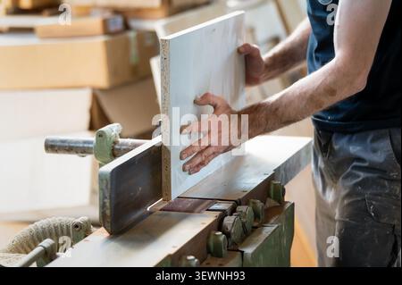 A carpenter guides a painted wood panel through a vertical edge sanding machine in a woodworking workshop, finishing the edges with precision using in Stock Photo