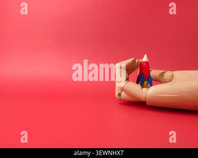 A wooden mannequin hand loosely cradles a small red, blue, and white toy rocket against a red background. Stock Photo
