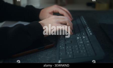 A close-up image showing hands typing on a computer keyboard with a smartphone resting next to it. Stock Photo