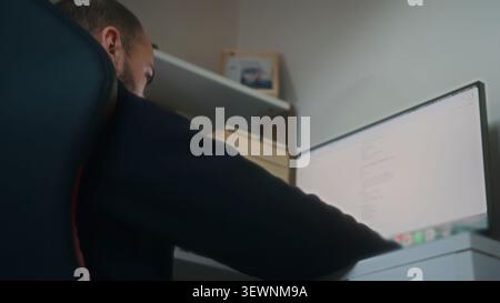 A man sits concentrated in a chair working on a computer screen in a cozy home office setting. Stock Photo