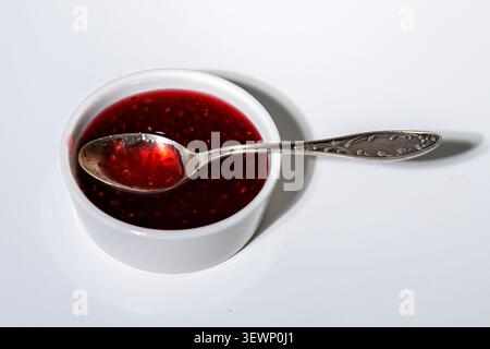 A white cup filled with vibrant raspberry jam. It shimmers in the light, revealing whole berries. Silver spoon. White background, close-up. Stock Photo