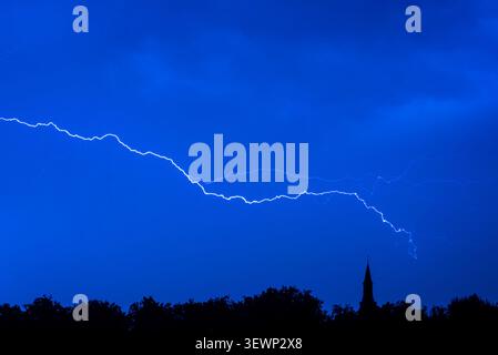Sheet lightning strike / intra-cloud lightning bolt during thunderstorm in blue night sky over forest with silhouetted treetops and church tower, Mari Stock Photo