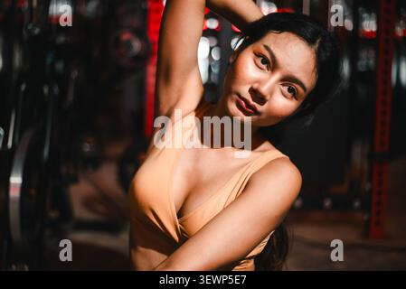 Close-up portrait of Asian woman doing side stretch in a dark gym, showing yoga flexibility, focused eyes, fitness training, body mobility, strength, Stock Photo
