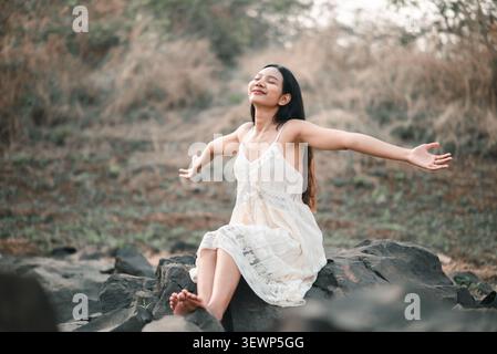 Asian woman in white lace dress sitting on rocks with arms open and eyes closed, enjoying fresh air, freedom, peaceful nature, relaxation, happiness, Stock Photo