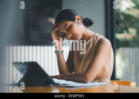 Concerned Asian businesswoman using tablet at cafe desk while reviewing financial paperwork, showing accounting pressure, budget analysis, digital wor Stock Photo
