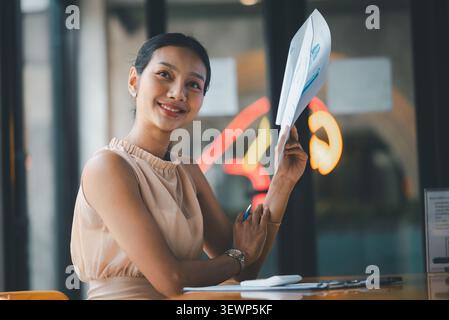 Smiling Asian businesswoman holding financial charts and pen in modern cafe office, presenting successful budget report, accounting analysis, corporat Stock Photo
