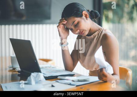 Stressed Asian businesswoman holding crumpled paper near laptop and financial charts, showing budget problems, accounting pressure, failed report, bus Stock Photo