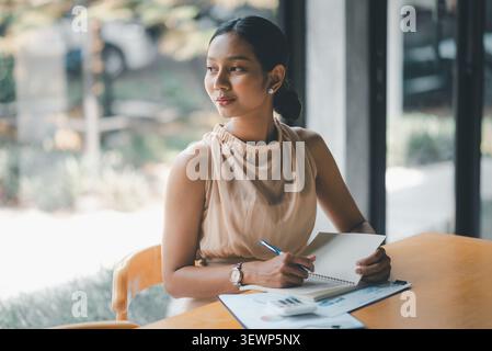 Thoughtful Asian businesswoman writing in notebook beside financial reports and calculator at cafe table, looking outside while planning budget, accou Stock Photo