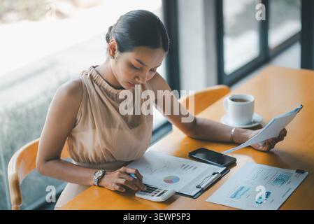 Top view of Asian businesswoman calculating financial data with reports, graphs, smartphone, and coffee on cafe table, ideal for budgeting, analytics, Stock Photo