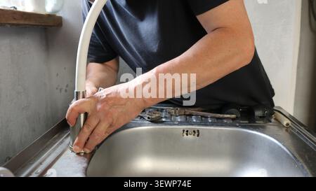 A man repairs a stainless steel kitchen faucet. His hands are holding the base of the faucet with a flexible white hose. The process of installing Stock Photo
