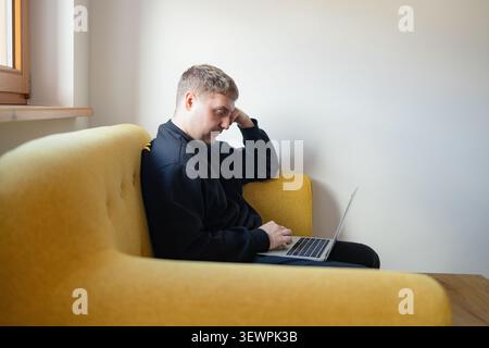 Thoughtful young man working on laptop on yellow sofa at home Stock Photo