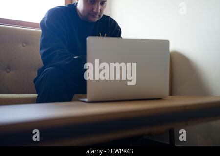 Thoughtful young man working on laptop on yellow sofa at home Stock Photo